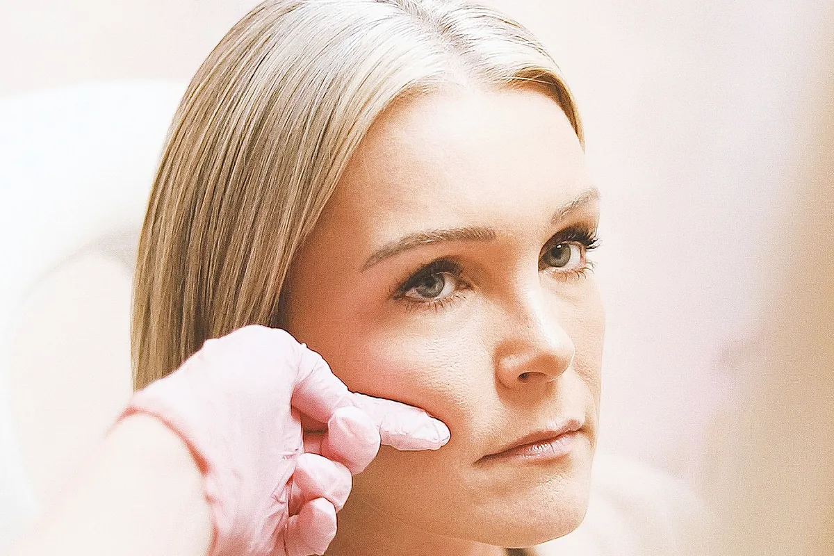 Behandlerin mit Handschuhen tastet vor der Injektion die Wange einer Kundin ab